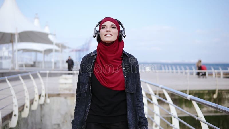 A menina muçulmana moderna com o hijab que cobre sua cabeça põe fones de ouvido sobre e começa-os andar em algum lugar apreciando vídeos de arquivo A menina muçulmana moderna com o hijab que cobre sua cabeça põe fones de ouvido sobre e começa-os andar em algum lugar apreciando vídeos de arquivo