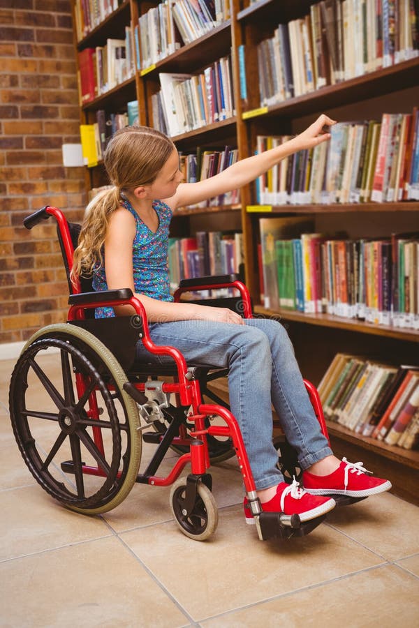 Menina na cadeira de rodas que seleciona o livro na biblioteca fotos de stock Menina na cadeira de rodas que seleciona o livro na biblioteca fotos de stock