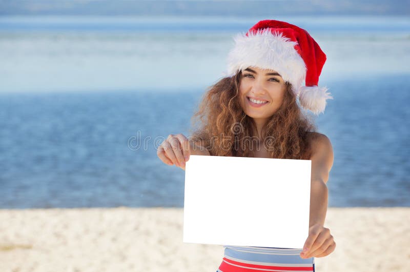 Menina nova, atrativa, encaracolado em um maiô e chapéu de Santa Claus na praia que guarda uma folha de papel branca imagem de stock royalty free Menina nova, atrativa, encaracolado em um maiô e chapéu de Santa Claus na praia que guarda uma folha de papel branca imagem de stock royalty free