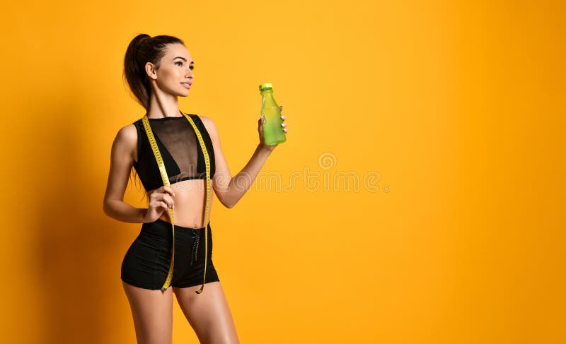 Cute young fitness girl in black shirt looks into the camera and keep the measuring tape and water at hand. Cute young fitness girl in black shirt looks into the camera and keep the measuring tape and water at hand.