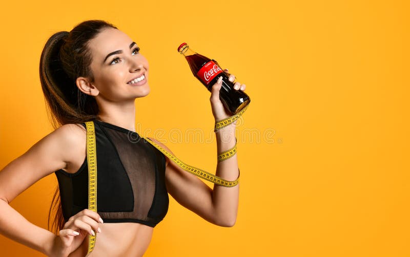 Moscow, Russia, January 2019 - cute young fitness girl in black sportswear looks into the camera and keep the measuring tape and water of Coca Cola at hand. Moscow, Russia, January 2019 - cute young fitness girl in black sportswear looks into the camera and keep the measuring tape and water of Coca Cola at hand