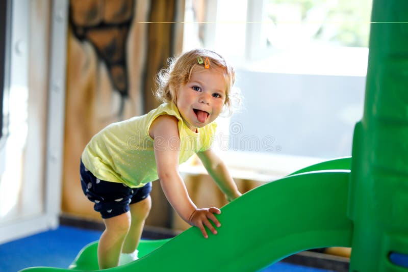 Menina pequena loura feliz da criança que tem o divertimento e que desliza no campo de jogos interno na guarda ou no berçário foto de stock royalty free Menina pequena loura feliz da criança que tem o divertimento e que desliza no campo de jogos interno na guarda ou no berçário foto de stock royalty free