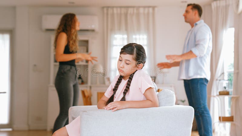 Annoyed and unhappy young girl sitting on sofa trapped in middle of tension by her parent argument in living room. Unhealthy domestic lifestyle and traumatic childhood develop to depression Synchronos. Annoyed and unhappy young girl sitting on sofa trapped in middle of tension by her parent argument in living room. Unhealthy domestic lifestyle and traumatic childhood develop to depression Synchronos