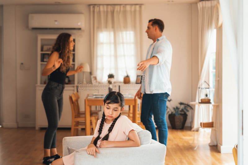 Annoyed and unhappy young girl sitting on sofa trapped in middle of tension by her parent argument in living room. Unhealthy domestic lifestyle and traumatic childhood develop to depression.Synchronos. Annoyed and unhappy young girl sitting on sofa trapped in middle of tension by her parent argument in living room. Unhealthy domestic lifestyle and traumatic childhood develop to depression.Synchronos