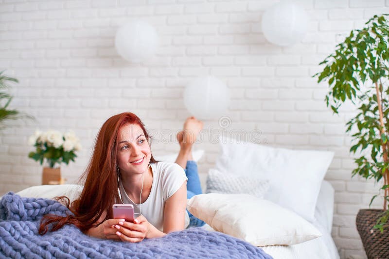 Menina que escuta a música nos fones de ouvido na cama luz brilhante da manhã do quarto das janelas fotografia de stock Menina que escuta a música nos fones de ouvido na cama luz brilhante da manhã do quarto das janelas fotografia de stock