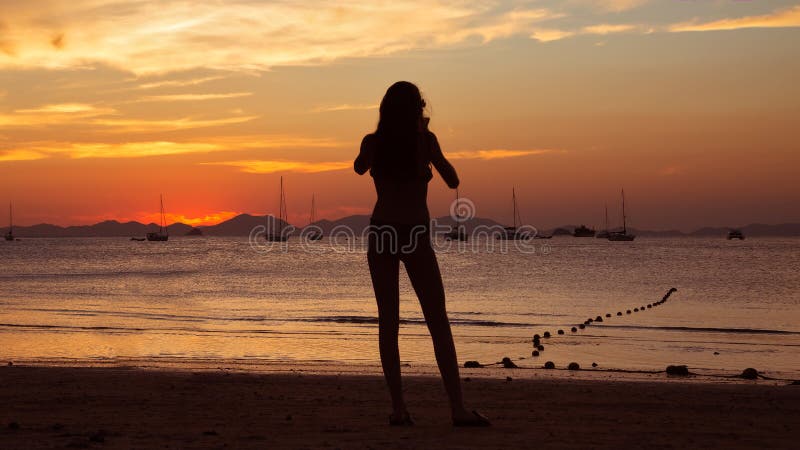 Menina que guarda o telefone esperto e que toma a foto vermelha do por do sol na praia tropical Silhueta de uma mulher, de iate e imagem de stock royalty free Menina que guarda o telefone esperto e que toma a foto vermelha do por do sol na praia tropical Silhueta de uma mulher, de iate e imagem de stock royalty free