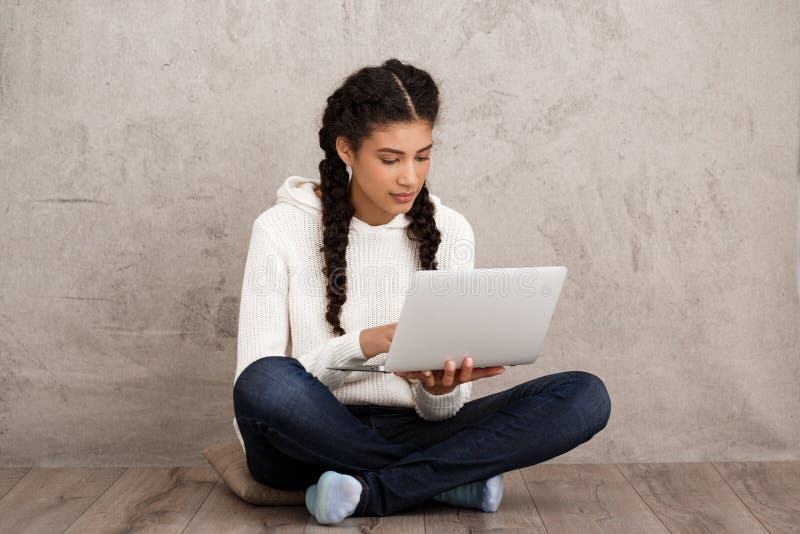 Menina que sorri, guardando o portátil, sentando-se no assoalho sobre o fundo bege imagem de stock Menina que sorri, guardando o portátil, sentando-se no assoalho sobre o fundo bege imagem de stock
