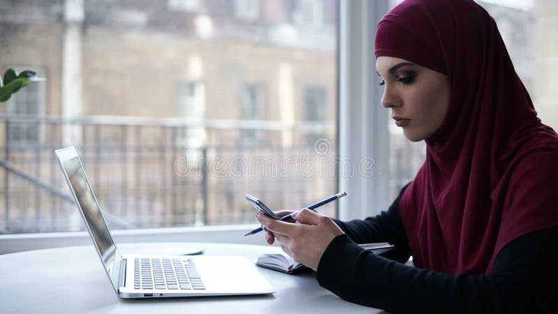 A menina supostamente muçulmana dos jovens com hijab roxo em sua cabeça escreve algo para baixo de seu smartphone, sentando-se em