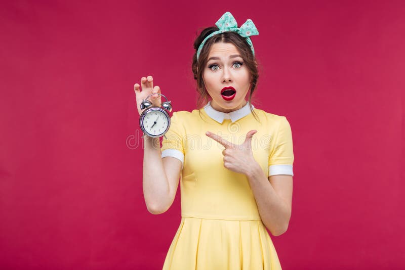 Menina surpreendida bonito do pinup que aponta no despertador imagem de stock Menina surpreendida bonito do pinup que aponta no despertador imagem de stock