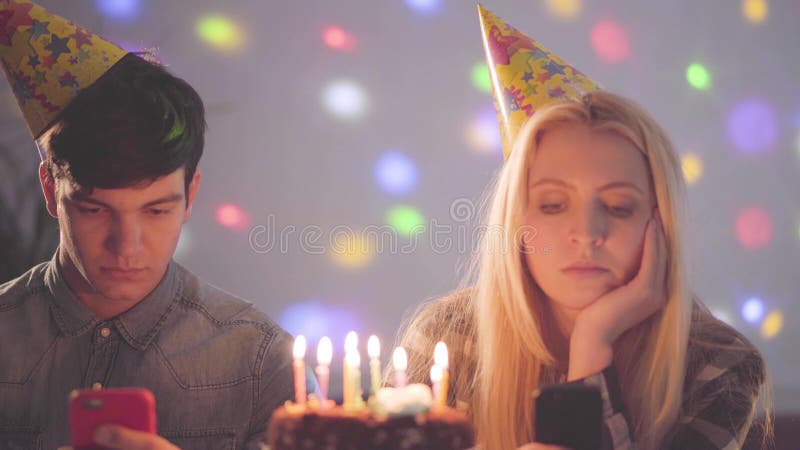 Menina triste do retrato e um homem nos chapéus do aniversário que sentam-se na frente do bolo com as velas que texting em seus t