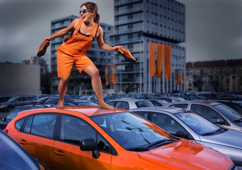 Menina à moda brincalhão nos macacões alaranjados que estão no telhado do carro no parque de estacionamento
