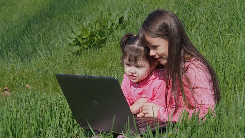 Meninas na grama verde com portátil Irmãs no parque em um dia ensolarado Meninas no rosa com um portátil Crianças no w