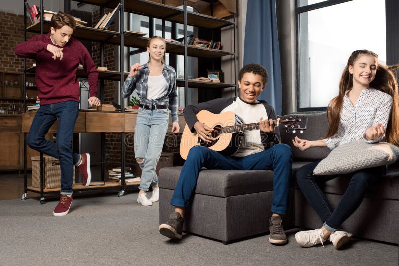 Menino afro-americano que joga a guitarra acustic quando seus amigos que dançam atrás em casa imagens de stock Menino afro-americano que joga a guitarra acustic quando seus amigos que dançam atrás em casa imagens de stock