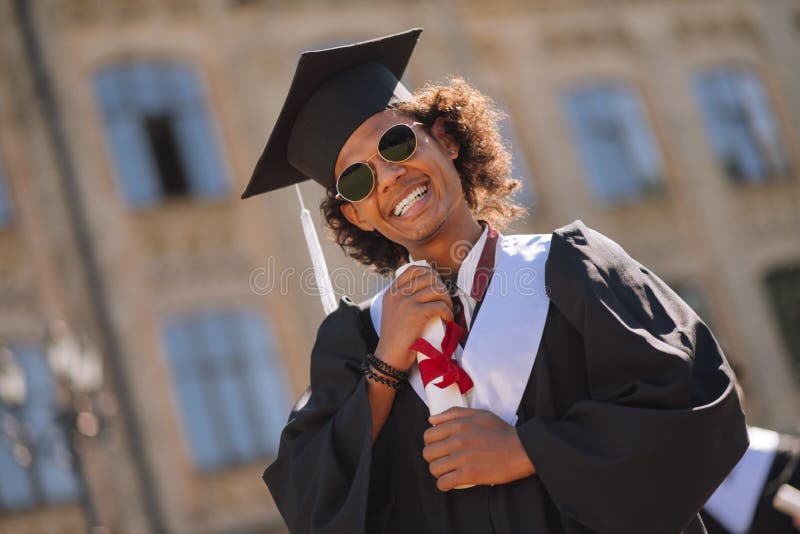 Menino alegre que abraça seu diploma na jarda de universidade imagem de stock royalty free Menino alegre que abraça seu diploma na jarda de universidade imagem de stock royalty free