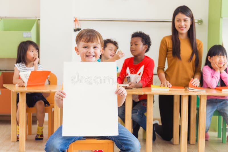 Menino bonito que guarda o cartaz branco vazio com a cara feliz na sala de aula do jardim de infância, conceito da educação do ja fotografia de stock Menino bonito que guarda o cartaz branco vazio com a cara feliz na sala de aula do jardim de infância, conceito da educação do ja fotografia de stock