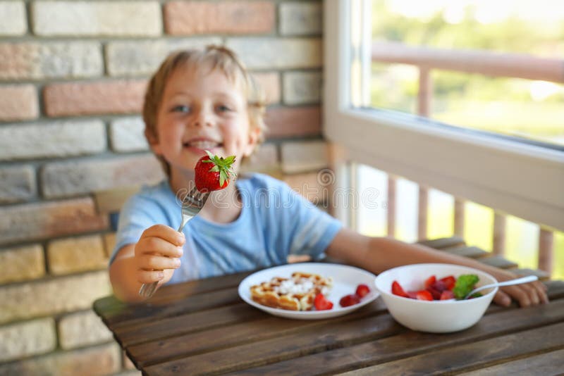 Menino caucasiano da criança de 5 anos feliz para comer para waffles vienenses do café da manhã com gelado e morangos