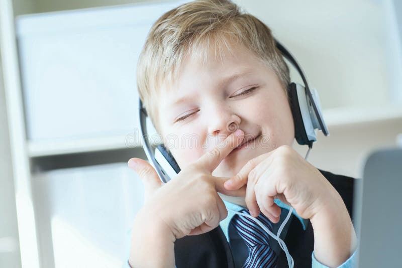 Menino feliz bonito da criança de 6 anos no terno que escuta a música ou o curso audio em fones de ouvido no fundo do escritório foto de stock Menino feliz bonito da criança de 6 anos no terno que escuta a música ou o curso audio em fones de ouvido no fundo do escritório foto de stock