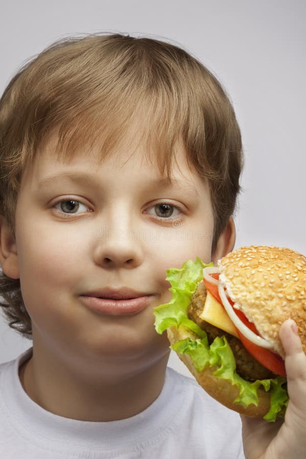 Menino feliz com hamburguer Adolescente com um sanduíche delicioso em um w imagem de stock royalty free Menino feliz com hamburguer Adolescente com um sanduíche delicioso em um w imagem de stock royalty free