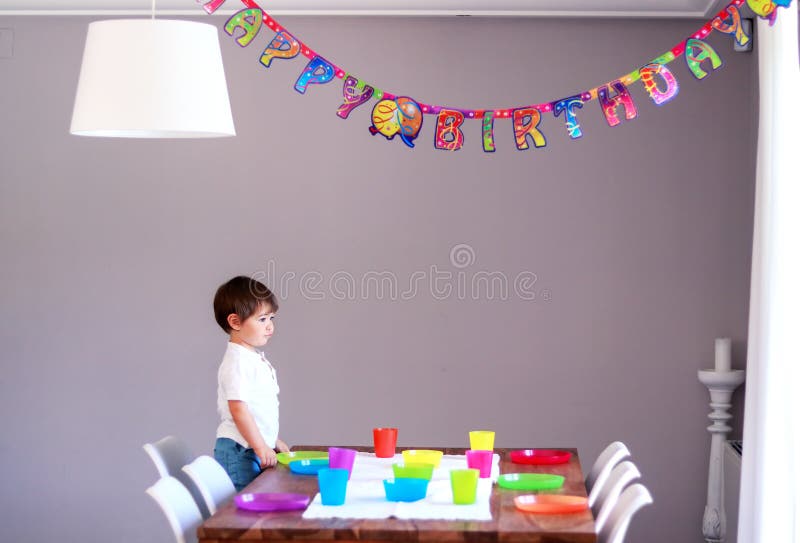 Menino thoghtful pequeno bonito que estabelece a tabela com a cutelaria colorida que prepara às crianças a festa de anos Decoraçã imagem de stock royalty free Menino thoghtful pequeno bonito que estabelece a tabela com a cutelaria colorida que prepara às crianças a festa de anos Decoraçã imagem de stock royalty free