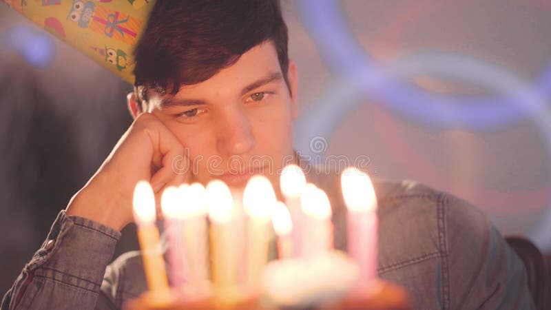 Menino triste só que senta-se na frente de pouco bolo com as velas iluminadas que olham nele O homem infeliz tem a festa de anos