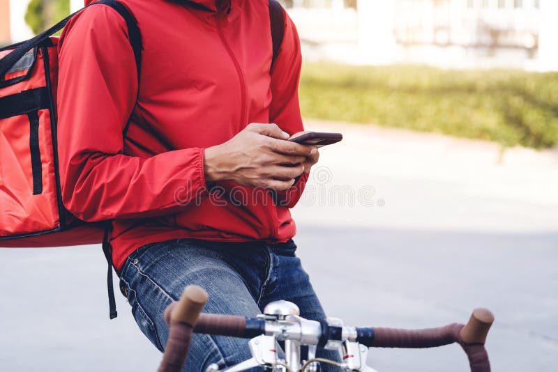 Mensageiro de uniforme vermelho com uma caixa de distribuição de volta andando de bicicleta e olhando para o telefone celular para