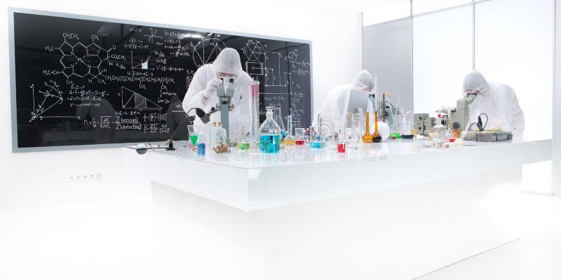 General-view of three people analysing substances in a chemistry lab using colorful liquids and lab tools with a blackboard with chemical formulas on the background. General-view of three people analysing substances in a chemistry lab using colorful liquids and lab tools with a blackboard with chemical formulas on the background