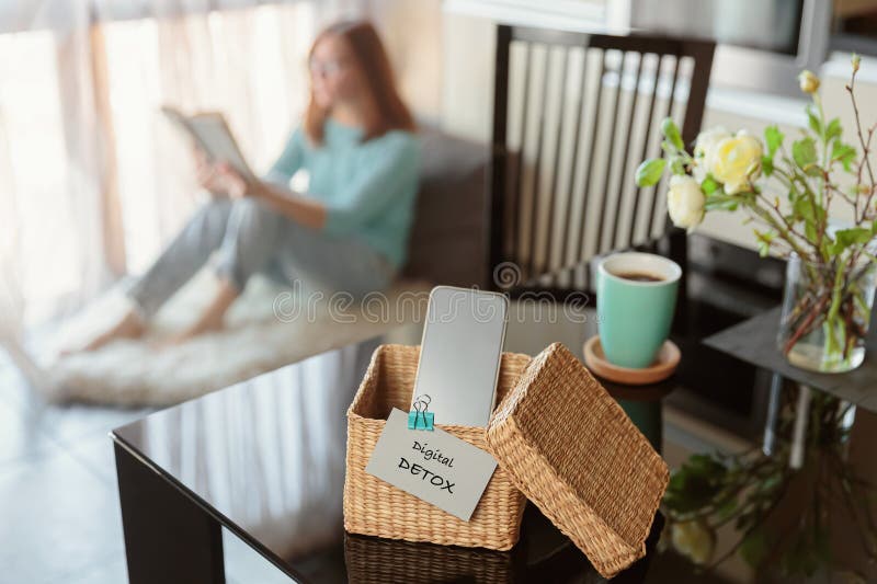 Smartphone is in separate wicker box with inscription digital detox on table. Woman reading book in background. Stop using digital gadgets. Mental and digital detox concept. Smartphone is in separate wicker box with inscription digital detox on table. Woman reading book in background. Stop using digital gadgets. Mental and digital detox concept