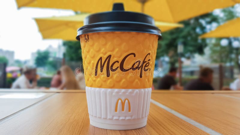 McCafe menu at McDonalds restaurant. Yellow cup of coffee on a table outdoors with yellow umbrellas and visitors in the background. Breakfast, coffee time. Ukraine, Kiev - September 6, 2019. McCafe menu at McDonalds restaurant. Yellow cup of coffee on a table outdoors with yellow umbrellas and visitors in the background. Breakfast, coffee time. Ukraine, Kiev - September 6, 2019.