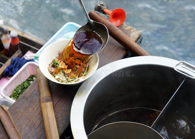 Merchant cook pouring herbal heat soup into white bowl of noodle thai style on floating wooden boat.