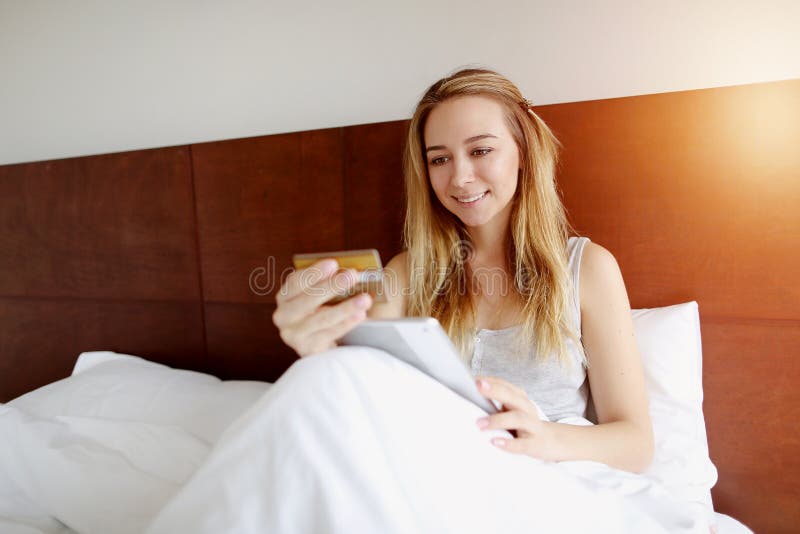 Merci d'acquisto della donna online soddisfatte della carta e della compressa di debito-credito che risiedono nel letto bianco a immagine stock libera da diritti Merci d'acquisto della donna online soddisfatte della carta e della compressa di debito-credito che risiedono nel letto bianco a immagine stock libera da diritti