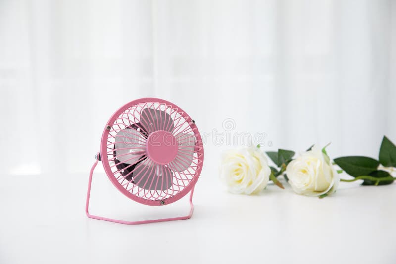 A pink small desk USB 4 blades cooling fan on a white table with roses in the background. A pink small desk USB 4 blades cooling fan on a white table with roses in the background