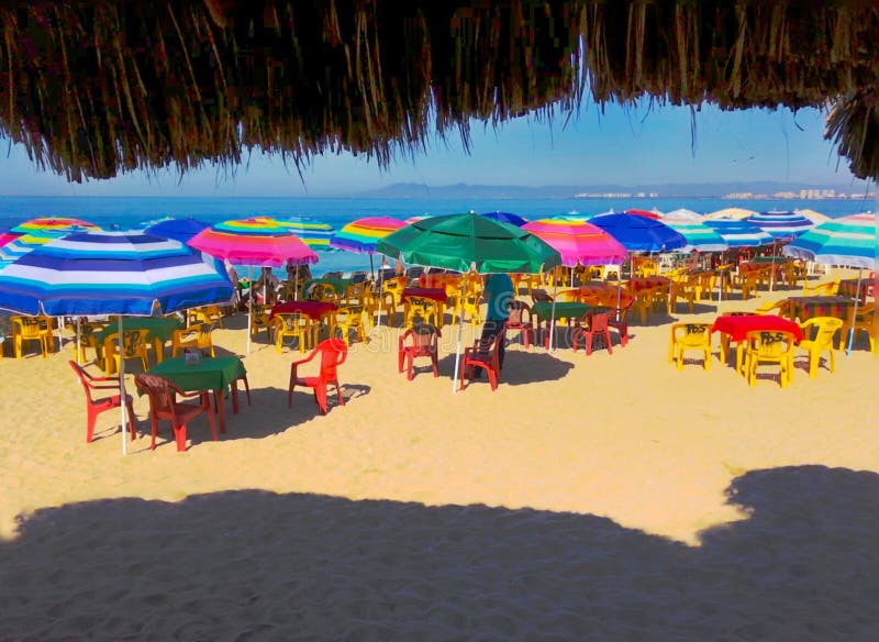 Pacific Ocean stark blue in the background of the beach scene in Puerto Vallarta, Mexico. Colorful umbrellas shade diners at restaurant tables from the hot sun. Fringe of palm leaves from the palapa frame the image. nThe palapa is very common on Mexican beaches and deserts. It is perhaps one of the most important architectural contributions of West Mexican culture. Pacific Ocean stark blue in the background of the beach scene in Puerto Vallarta, Mexico. Colorful umbrellas shade diners at restaurant tables from the hot sun. Fringe of palm leaves from the palapa frame the image. nThe palapa is very common on Mexican beaches and deserts. It is perhaps one of the most important architectural contributions of West Mexican culture.