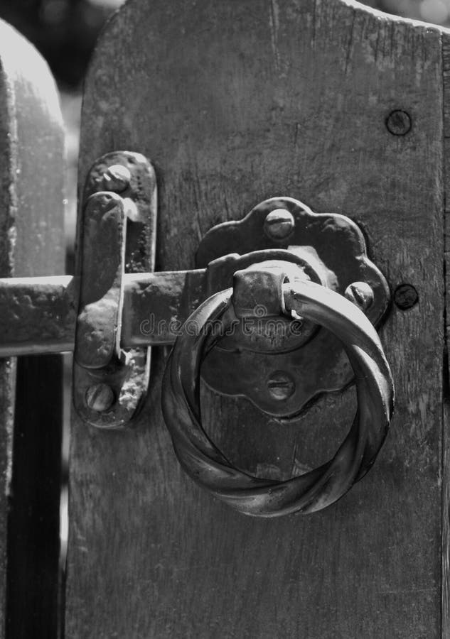 Ornate metal latch with a twisted ring handle is mounted on a wooden gate. The latch features a decorative backplate with visible screws, showcasing a combination of rustic and elegant design elements. The wood is dark and appears weathered, adding to the vintage aesthetic. The latch's metallic elements show signs of wear, indicating age and use. The contrasting textures of metal and wood create a visually appealing combination in this close-up view. Elegant lighting signs stock images, royalty-free photos and pictures