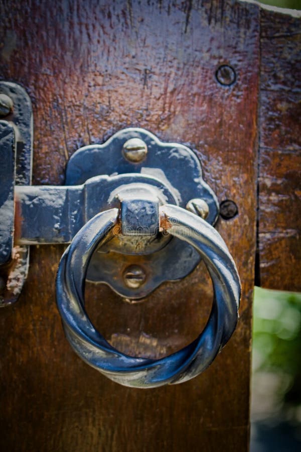 Ornate metal latch with a twisted ring handle is mounted on a wooden gate. The latch features a decorative backplate with visible screws, showcasing a combination of rustic and elegant design elements. The wood is dark and appears weathered, adding to the vintage aesthetic. The latch's metallic elements show signs of wear, indicating age and use. The contrasting textures of metal and wood create a visually appealing combination in this close-up view. Elegant lighting signs stock images, royalty-free photos and pictures