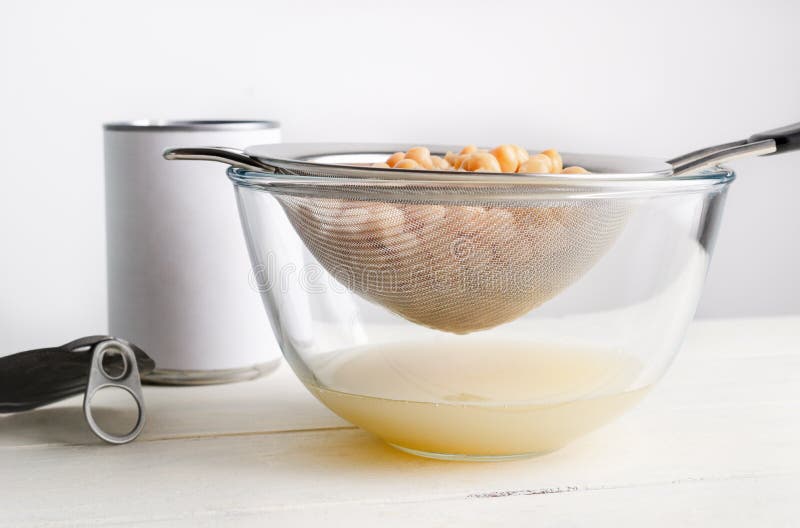 Metal sieve draining aquafaba chickpea water into glass bowl. Unbranded open tin with lid resting to left on wood plank table. Eye level shot. Metal sieve draining aquafaba chickpea water into glass bowl. Unbranded open tin with lid resting to left on wood plank table. Eye level shot