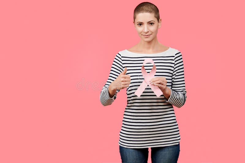 Metà di 30s donna sorridente, un superstite del cancro, tenendo il nastro rosa di consapevolezza del cancro al seno e mostrando i fotografie stock libere da diritti Metà di 30s donna sorridente, un superstite del cancro, tenendo il nastro rosa di consapevolezza del cancro al seno e mostrando i fotografie stock libere da diritti