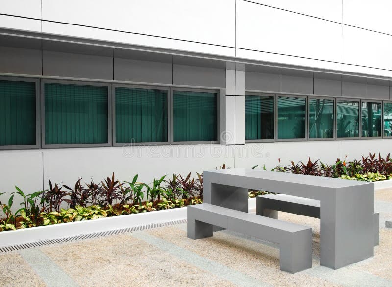 A photograph showing the court yard garden of a modern office building, with newly planted landscaping shrubs, and modern simple furniture of table and benches. A photograph showing the court yard garden of a modern office building, with newly planted landscaping shrubs, and modern simple furniture of table and benches.