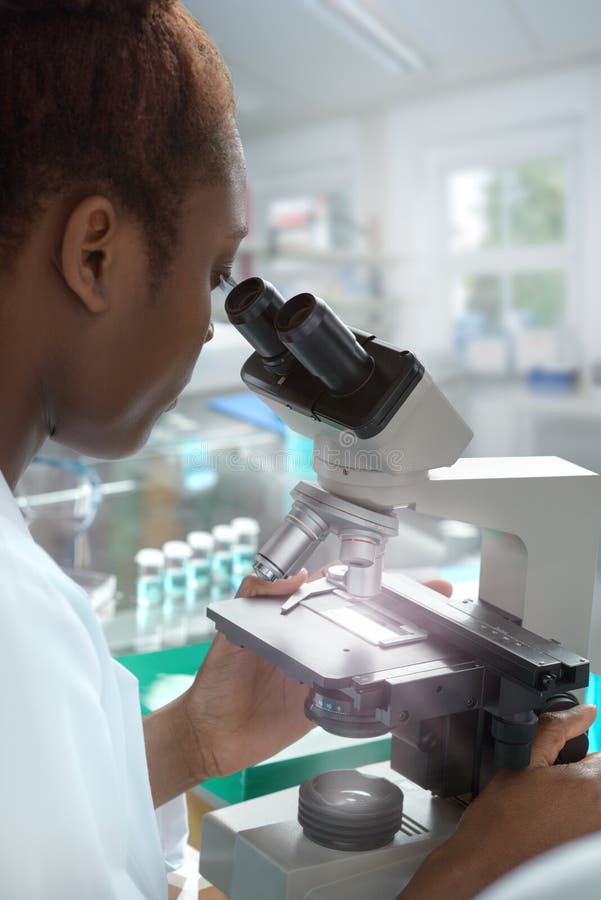 African-american female scientist or graduate student in lab coat works with a microscope in modern laboratory. African-american female scientist or graduate student in lab coat works with a microscope in modern laboratory