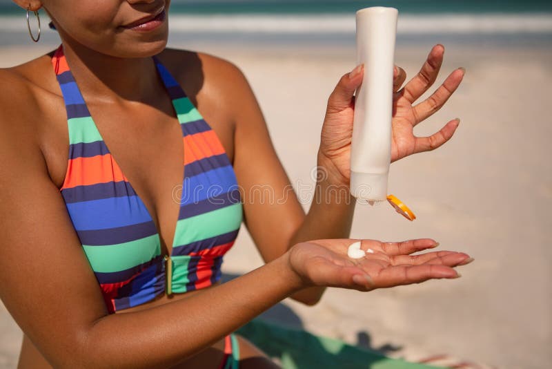 Mid section of African american woman in bikini applying sunscreen lotion at beach in the sunshine