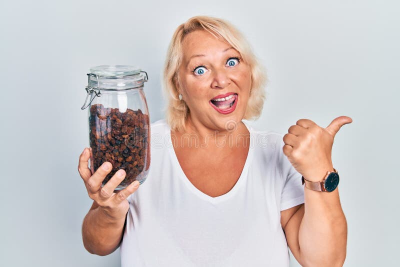 Middle age blonde woman holding raisins bowl pointing thumb up to the side smiling happy with open mouth stock image