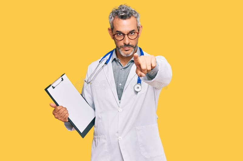 Middle age grey-haired man wearing doctor stethoscope holding clipboard pointing with finger to the camera and to you, confident gesture looking serious