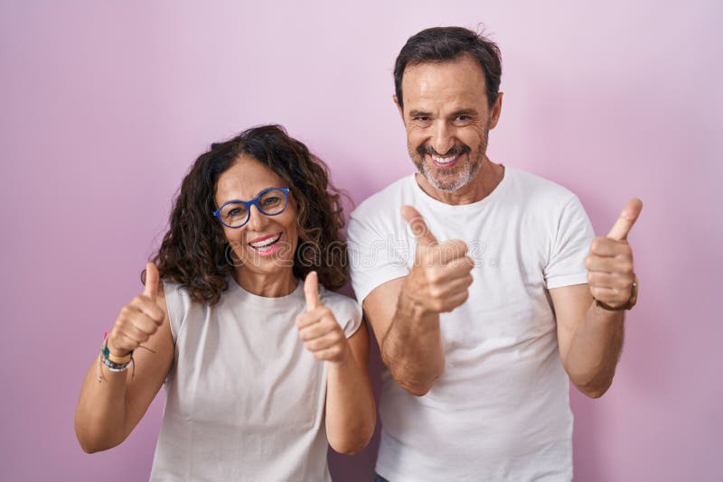Middle age hispanic couple together over pink background success sign doing positive gesture with hand, thumbs up smiling and