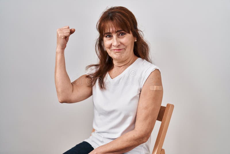 Middle age hispanic woman getting vaccine showing arm with band aid strong person showing arm muscle, confident and proud of power