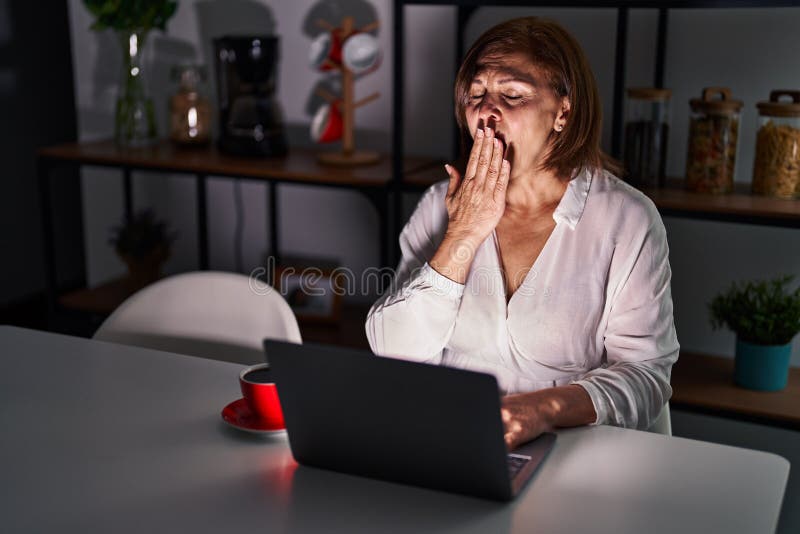 Middle age hispanic woman using laptop at home at night bored yawning tired covering mouth with hand