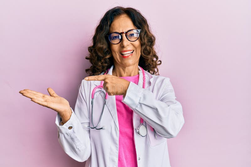 Middle age hispanic woman wearing doctor uniform and glasses amazed and smiling to the camera while presenting with hand and. Pointing with finger