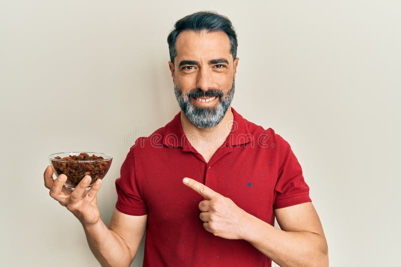 Middle age man with beard and grey hair holding raisins bowl smiling happy pointing with hand and finger stock photo