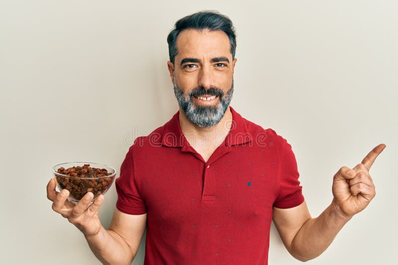 Middle age man with beard and grey hair holding raisins bowl smiling happy pointing with hand and finger to the side stock photos