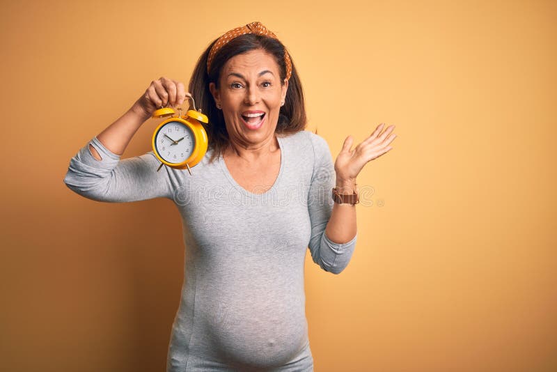Middle age pregnant woman expecting baby at aged pregnancy holding alarm clock very happy and excited, winner expression celebrating victory screaming with big smile and raised hands