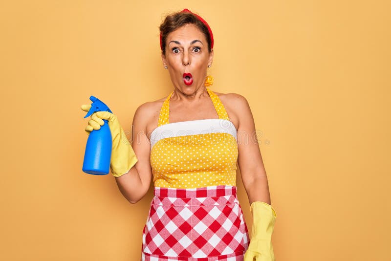 Middle age senior housewife pin up woman wearing 50s style retro dress cleaning using spray scared in shock with a surprise face, afraid and excited with fear expression