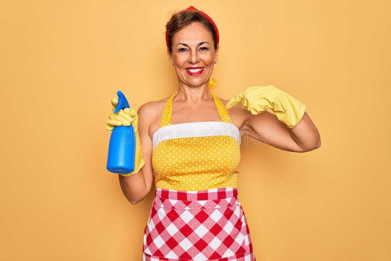 Middle age senior housewife pin up woman wearing 50s style retro dress cleaning using spray with surprise face pointing finger to. Himself